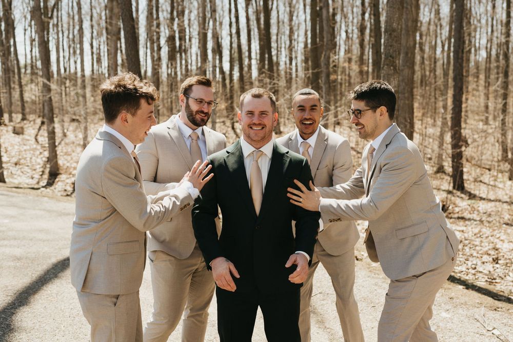 Groom in black suit laughing with four groomsmen in tan suits outdoors among bare trees on wedding day.