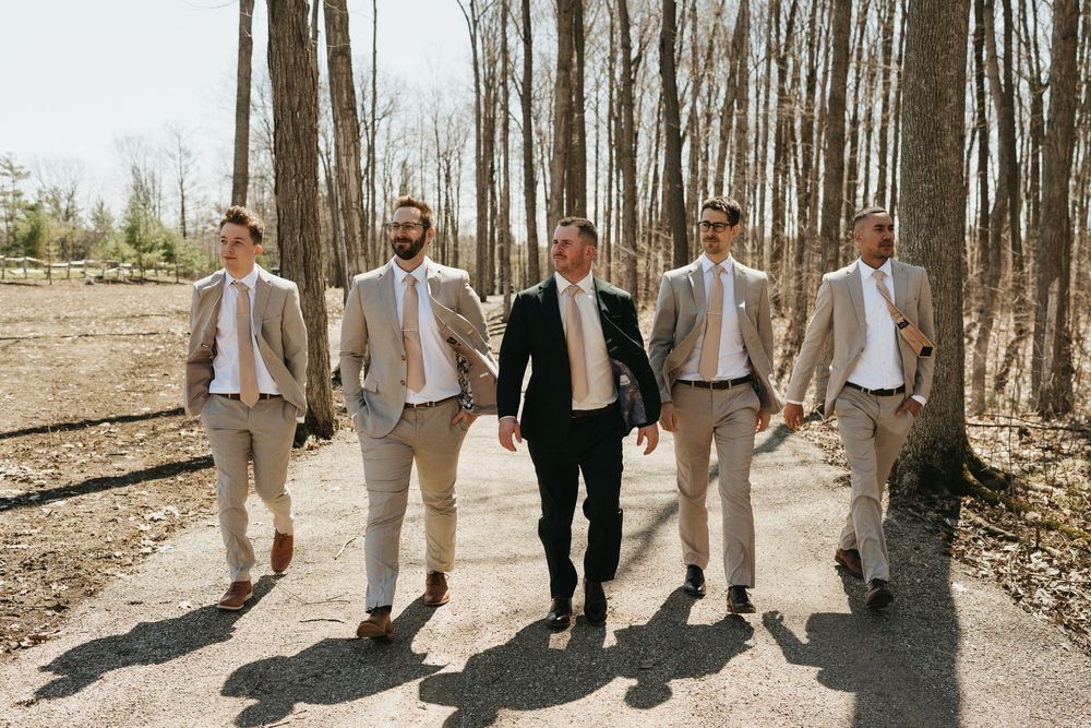 Groom and groomsmen in tan suits walk together on a sunlit forest path at an outdoor wedding.