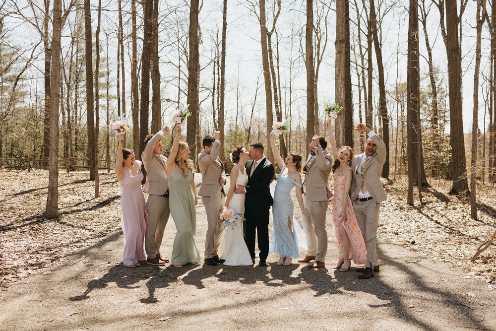 Wedding party celebrating outdoors in a wooded setting, bridesmaids in mixed pastel gowns and groomsmen in tan suits.