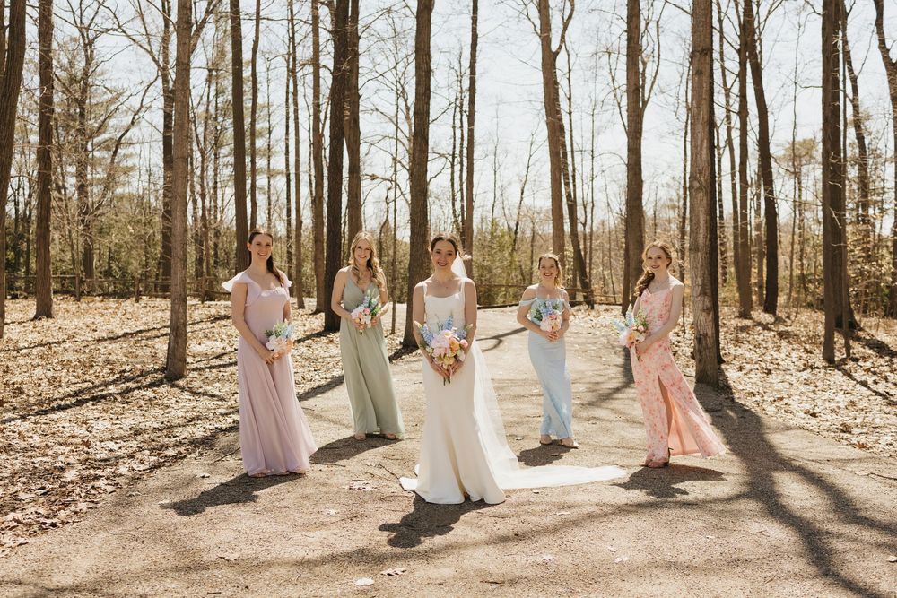 Bride and bridesmaids in pastel gowns pose together on a wooded path holding bouquets.