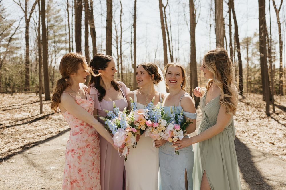 Bride and four bridesmaids in colorful dresses holding bouquets laughing together in a sunlit wooded outdoor setting.