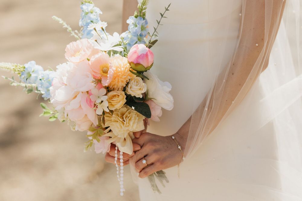 Bride holds a pastel bouquet of pink peonies, blue delphiniums, and yellow roses on wedding day.