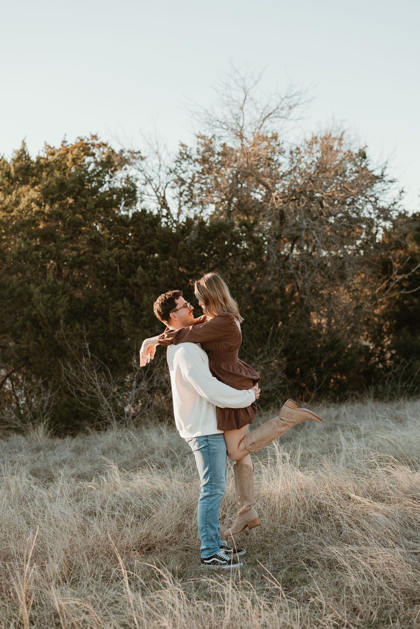 Couple holding hands and walking through a wooded park, woman in floral maxi dress, man in beige sweater.
