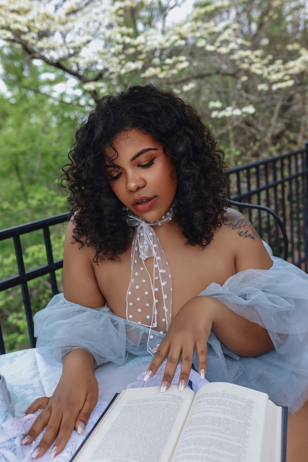 A curly-haired woman in a sheer blue off-shoulder gown reads a book on an outdoor balcony surrounded by blooming trees.