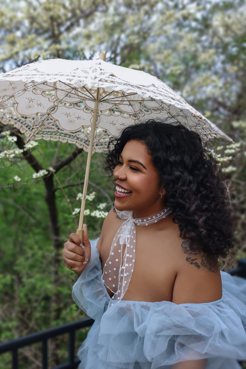 Smiling woman with curly hair holding lace parasol wearing off-shoulder blue tulle dress outdoors.