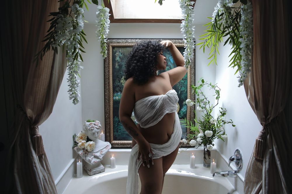 A curvy woman draped in sheer white fabric poses elegantly in a decorated bathroom with lush greenery and floral arrangements.