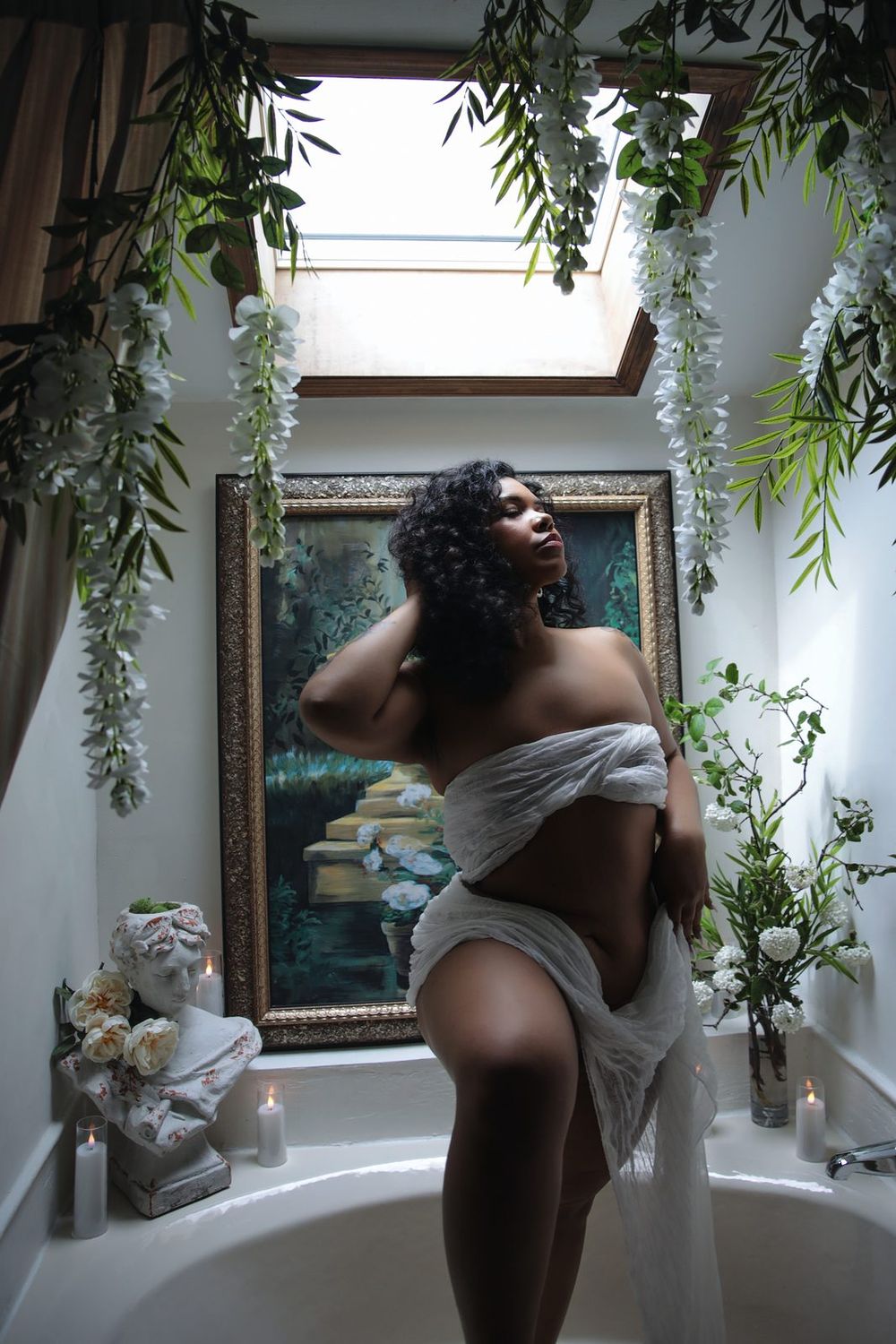 Woman draped in white fabric poses elegantly in a floral bathroom with candles, greenery, and a classical painting.