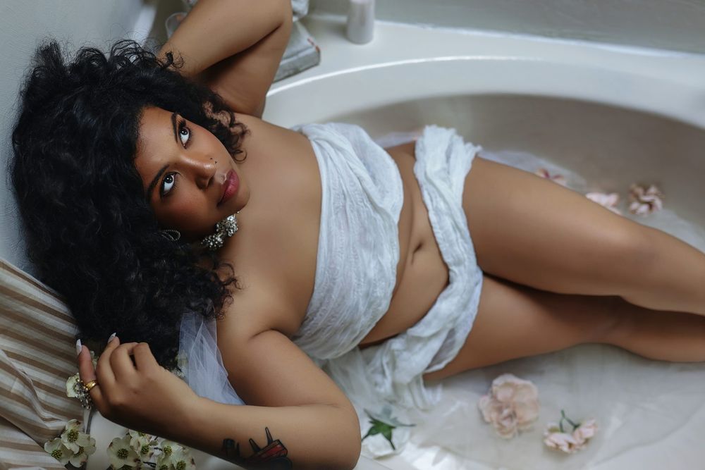 Woman with curly hair reclining in bathtub with white fabric and scattered rose petals.