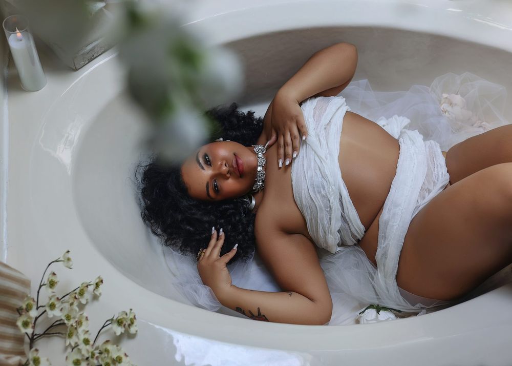 A woman in white fabric reclines gracefully in a bathtub, smiling softly in an elegant boudoir-style photo.