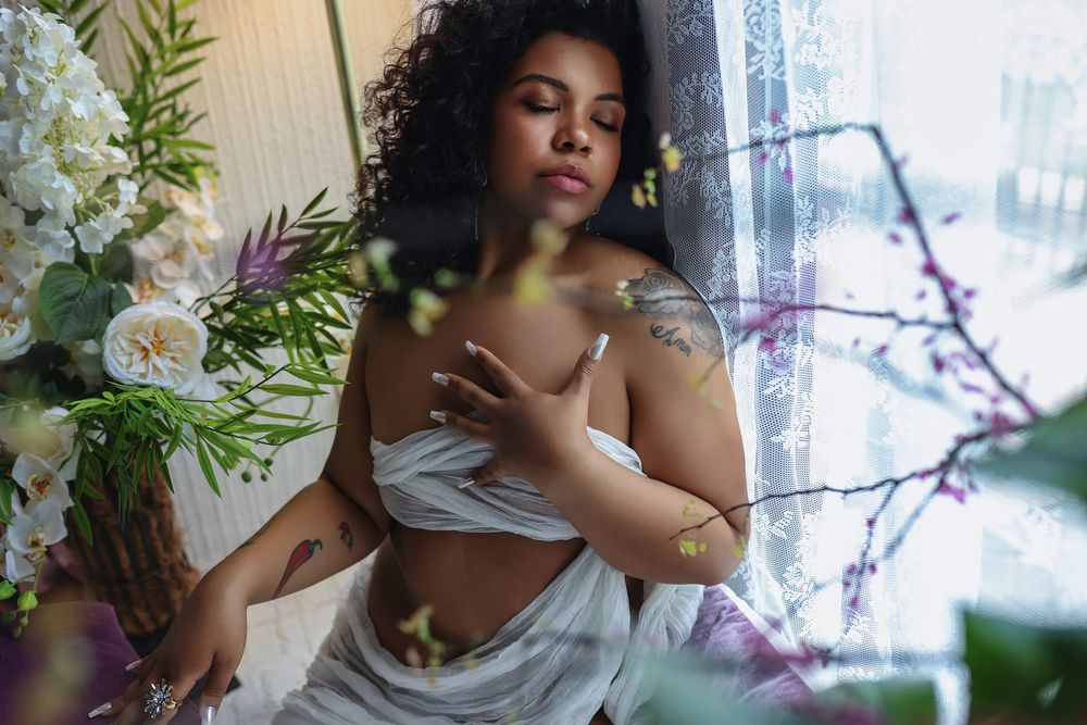 A woman with curly hair poses gracefully among flowers near a window, wearing a white wrap, showcasing tattoos on her arms.