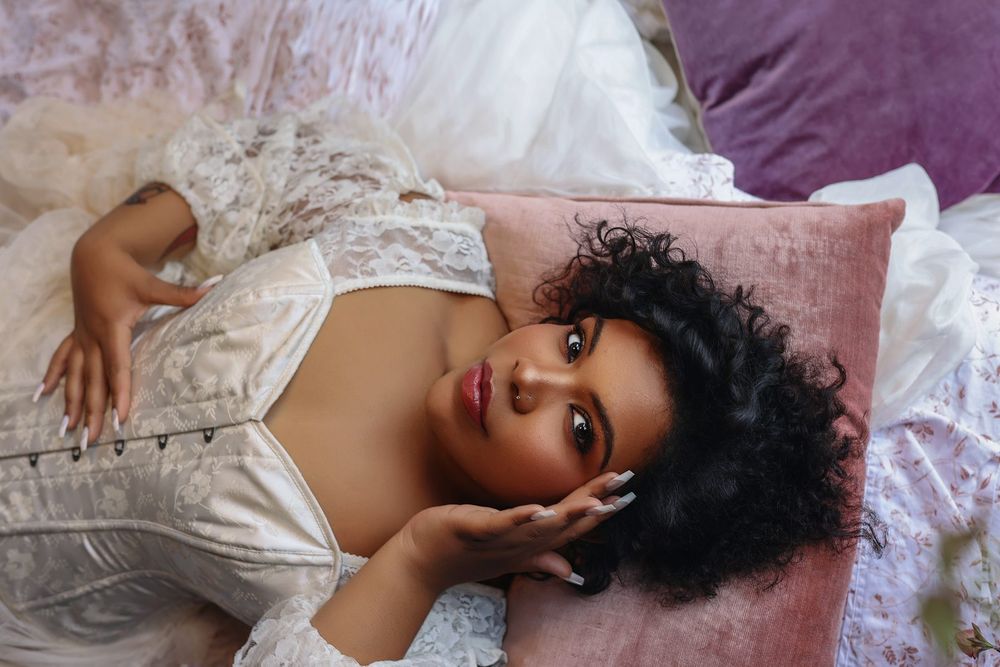 A young woman with curly hair in white lace reclines gracefully on a pink velvet pillow.