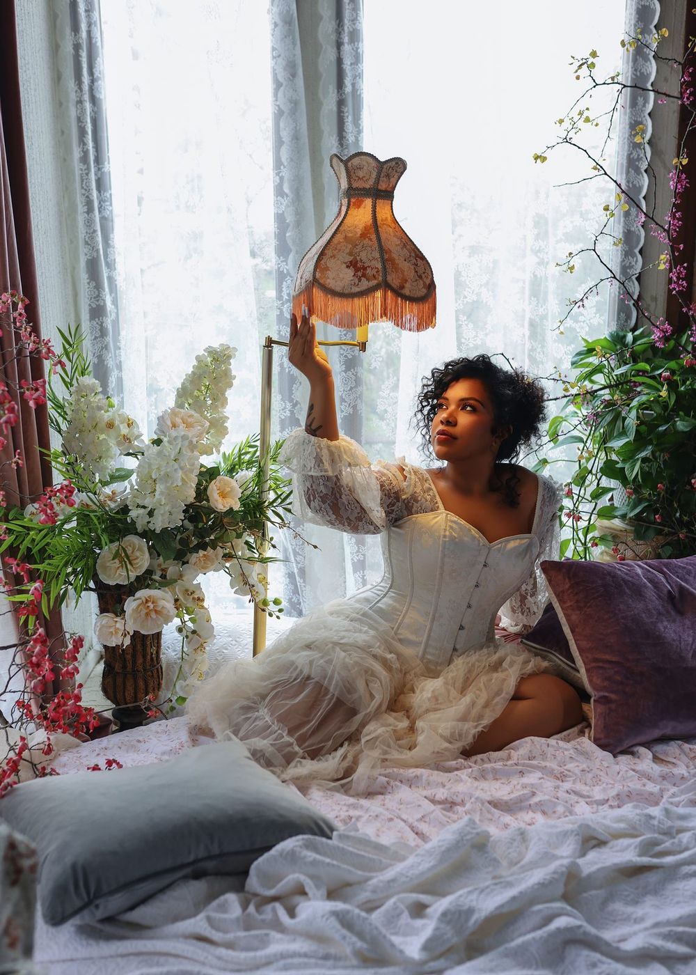 Woman in white corset and tulle skirt holds vintage lamp while sitting among flowers by a window.
