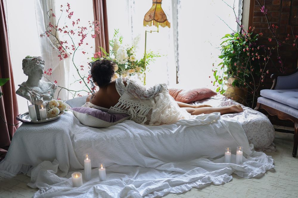 Woman relaxing on a boho-style bed with white linens, candles, plants, and decorative pillows in a cozy room.