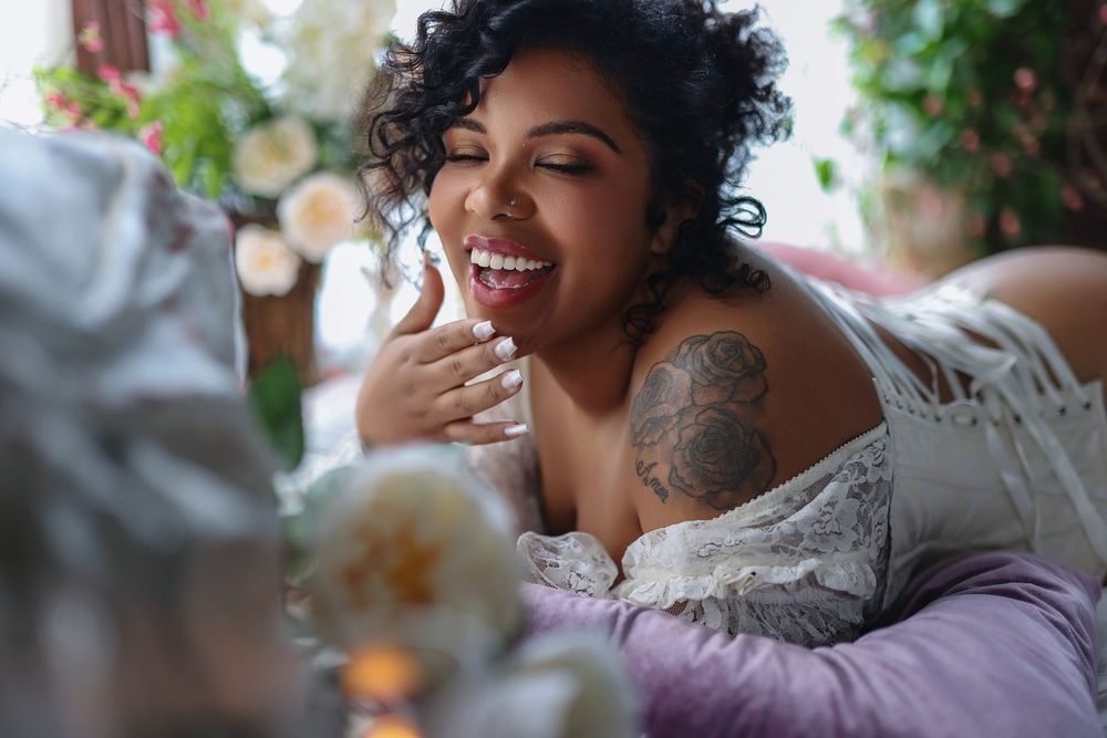 A joyful plus-size woman with curly hair and a shoulder tattoo laughs while wearing an off-shoulder top near white flowers.