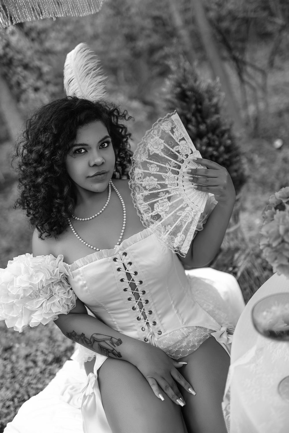 Black and white photo of a woman in a white corset holding a lace fan outdoors.