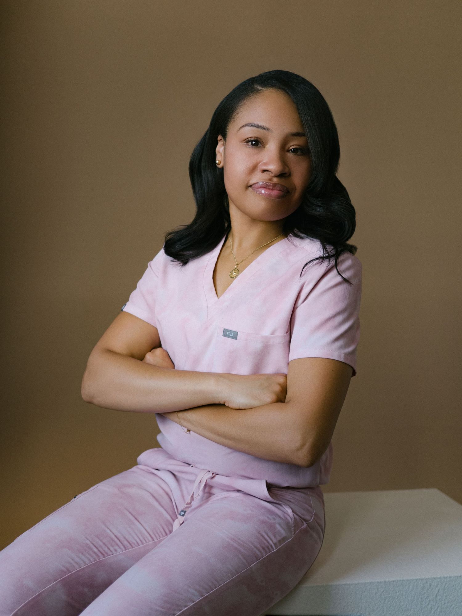 A healthcare professional in pink scrubs sits confidently with arms crossed against a warm brown background.