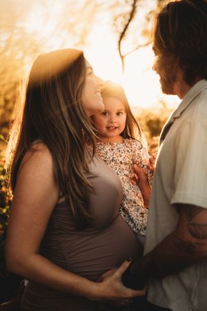 Pregnant woman and man holding toddler girl during golden hour outdoor maternity photo session.