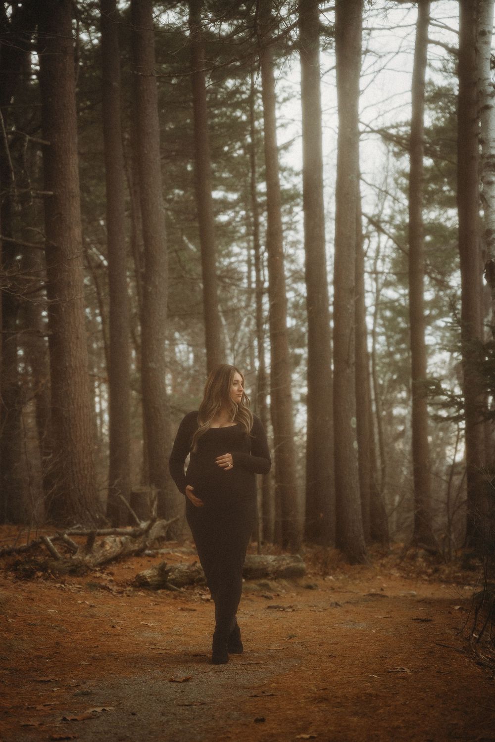 A woman in black walks a misty forest path surrounded by tall trees with warm golden backlight filtering through.