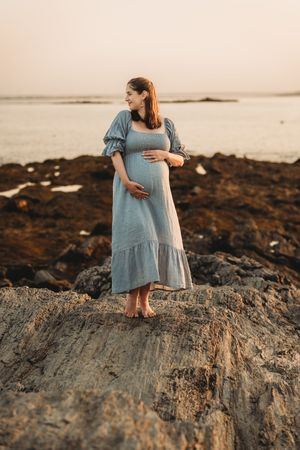 Pregnant woman in blue dress standing on rocky beach at sunset, cradling her baby bump with ocean in background.