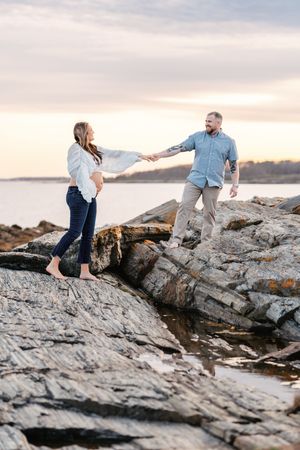 Couple holding hands on rocky coastal shore at sunset, woman in white blouse and navy pants, man in blue shirt.