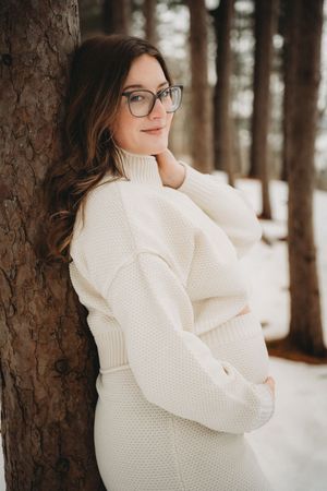 Woman in white knit sweater dress leaning against tree in snowy forest, wearing glasses, smiling softly.