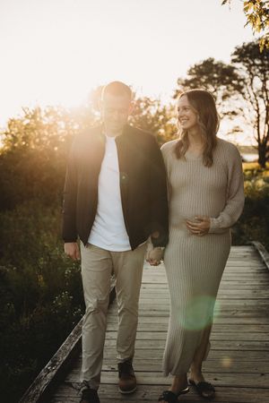 Couple walks hand-in-hand on wooden boardwalk at golden sunset, woman in beige knit dress cradling baby bump.