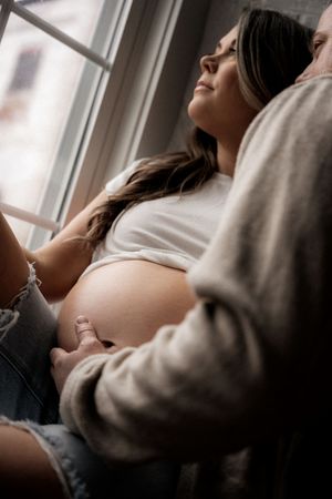 Pregnant woman in white top sits by window, partner tenderly touching her bare baby bump.