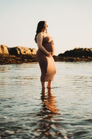 Pregnant woman in tan dress standing in shallow ocean water at golden hour with rocky shore backdrop.