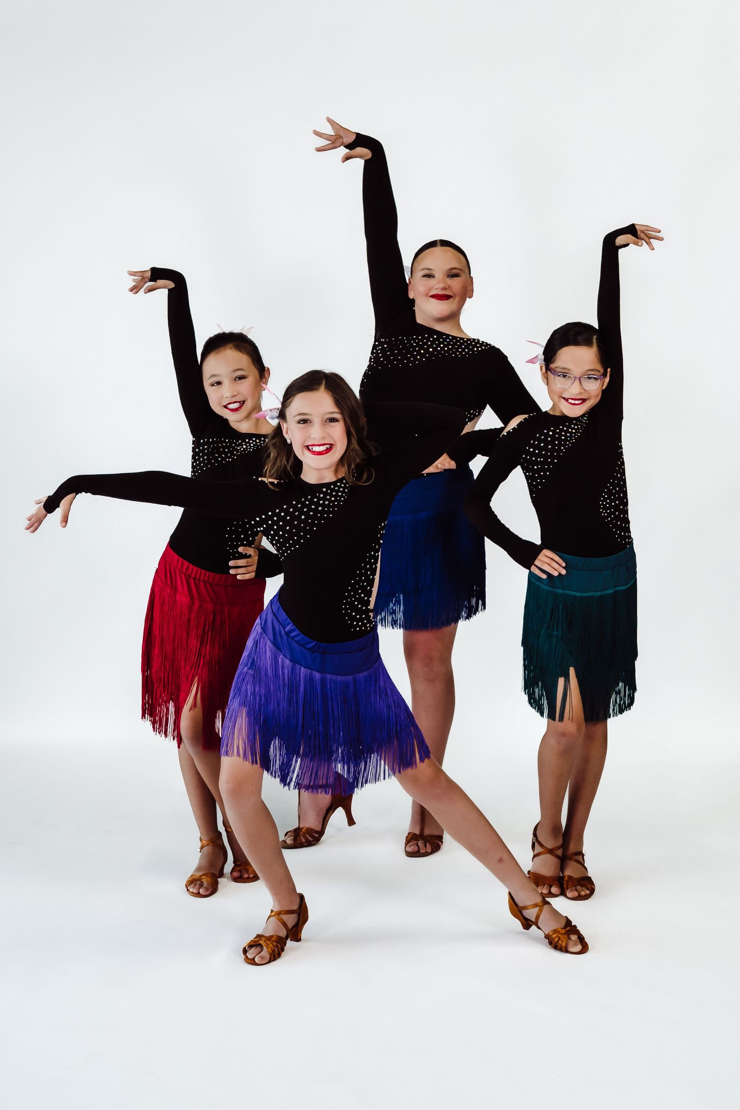Four young girls in black rhinestone tops and colorful fringe skirts pose in Latin dance formation against white background.