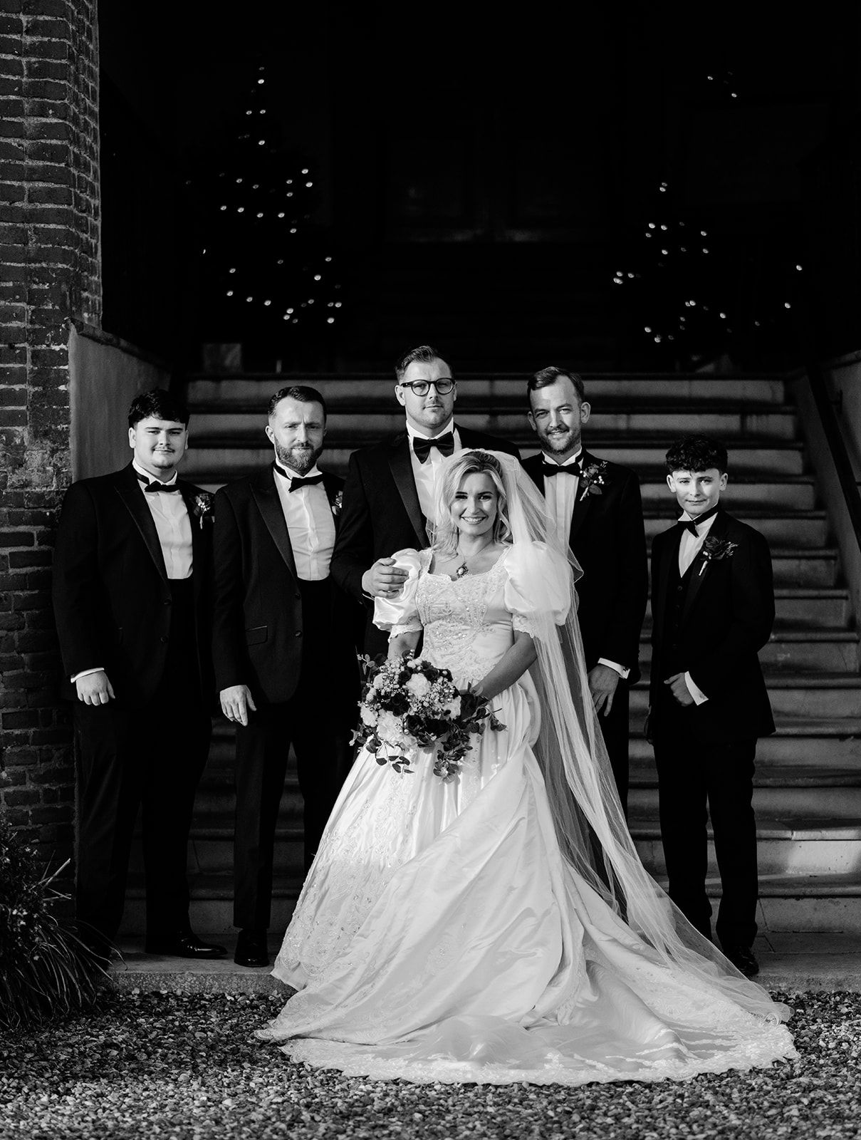 Black and white wedding photo of bride in gown with veil holding bouquet, surrounded by groom and groomsmen on steps.