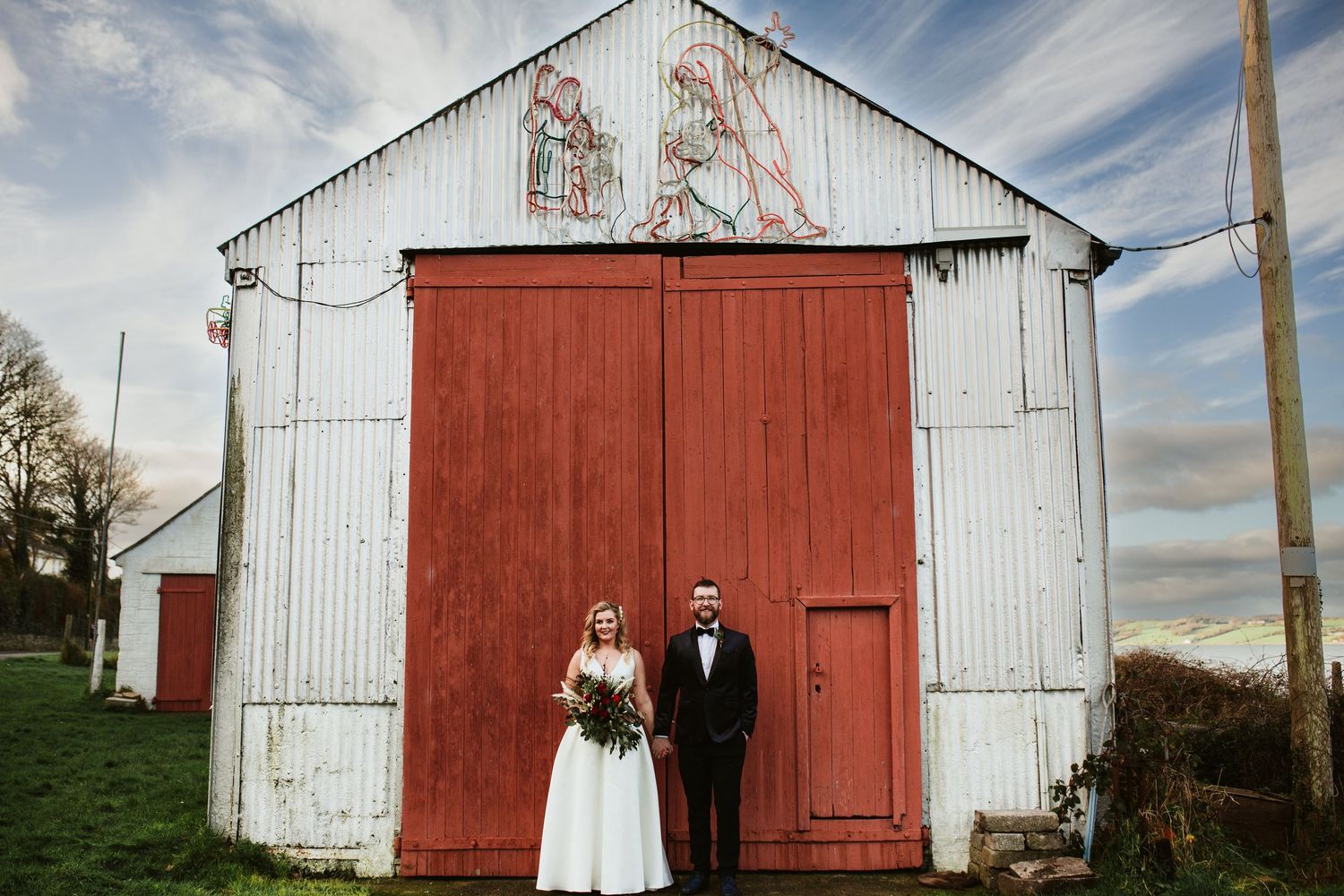 Bride and groom standing before a rustic white barn with red doors on their wedding day.