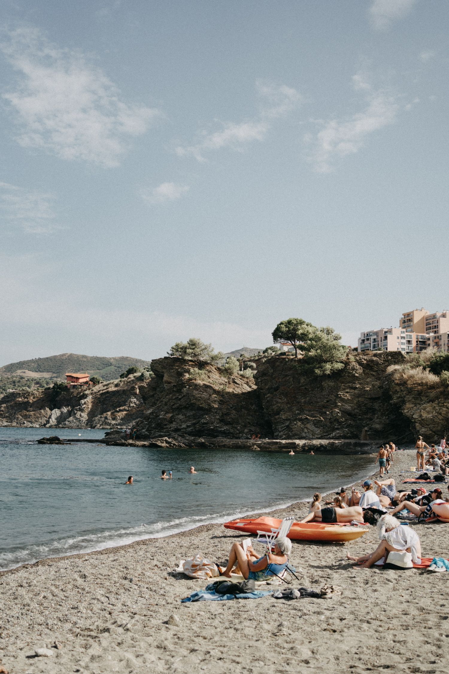 Sunbathers relax on a rocky Mediterranean beach with kayaks, swimmers, and a scenic coastal cliff backdrop.