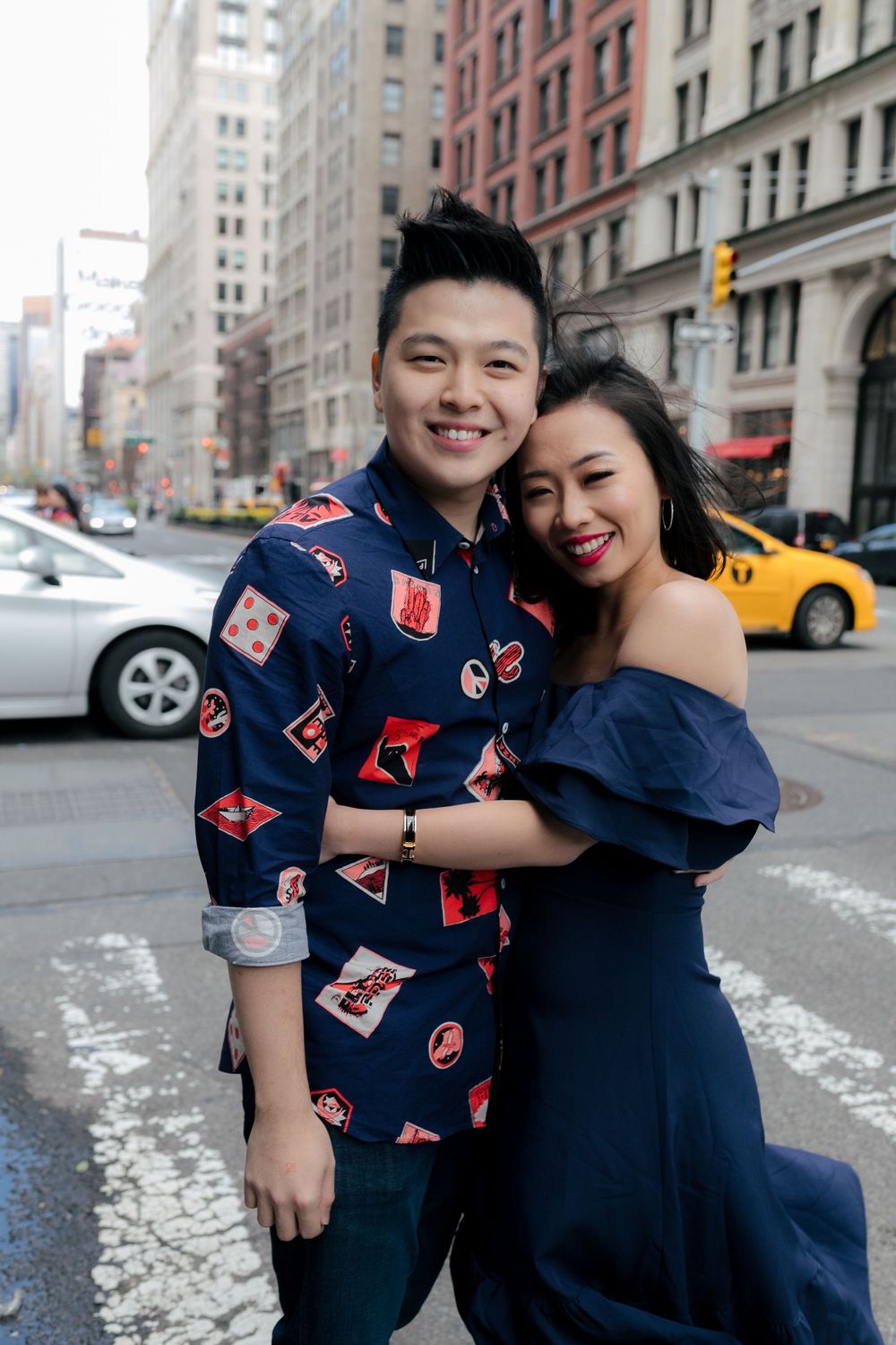 Smiling Asian couple embracing on a New York City street, man in patterned shirt, woman in navy off-shoulder dress.