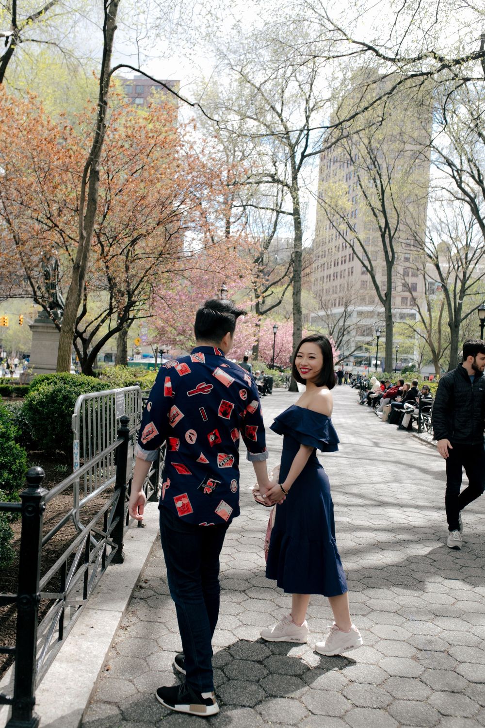 Couple holding hands walking along a tree-lined path in a city park during spring bloom.