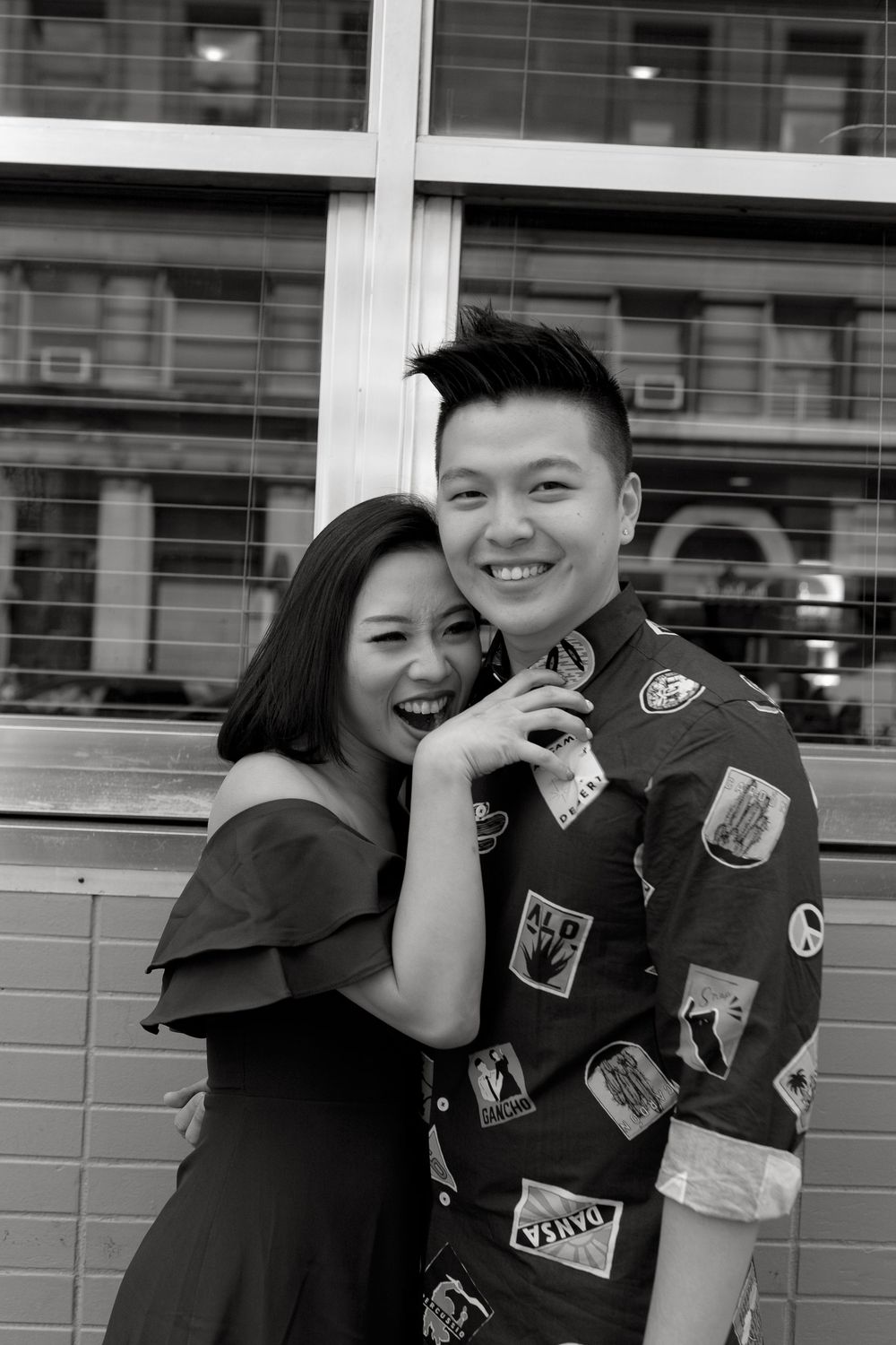 Joyful Asian couple posing together in black and white, woman in off-shoulder dress hugging smiling man.