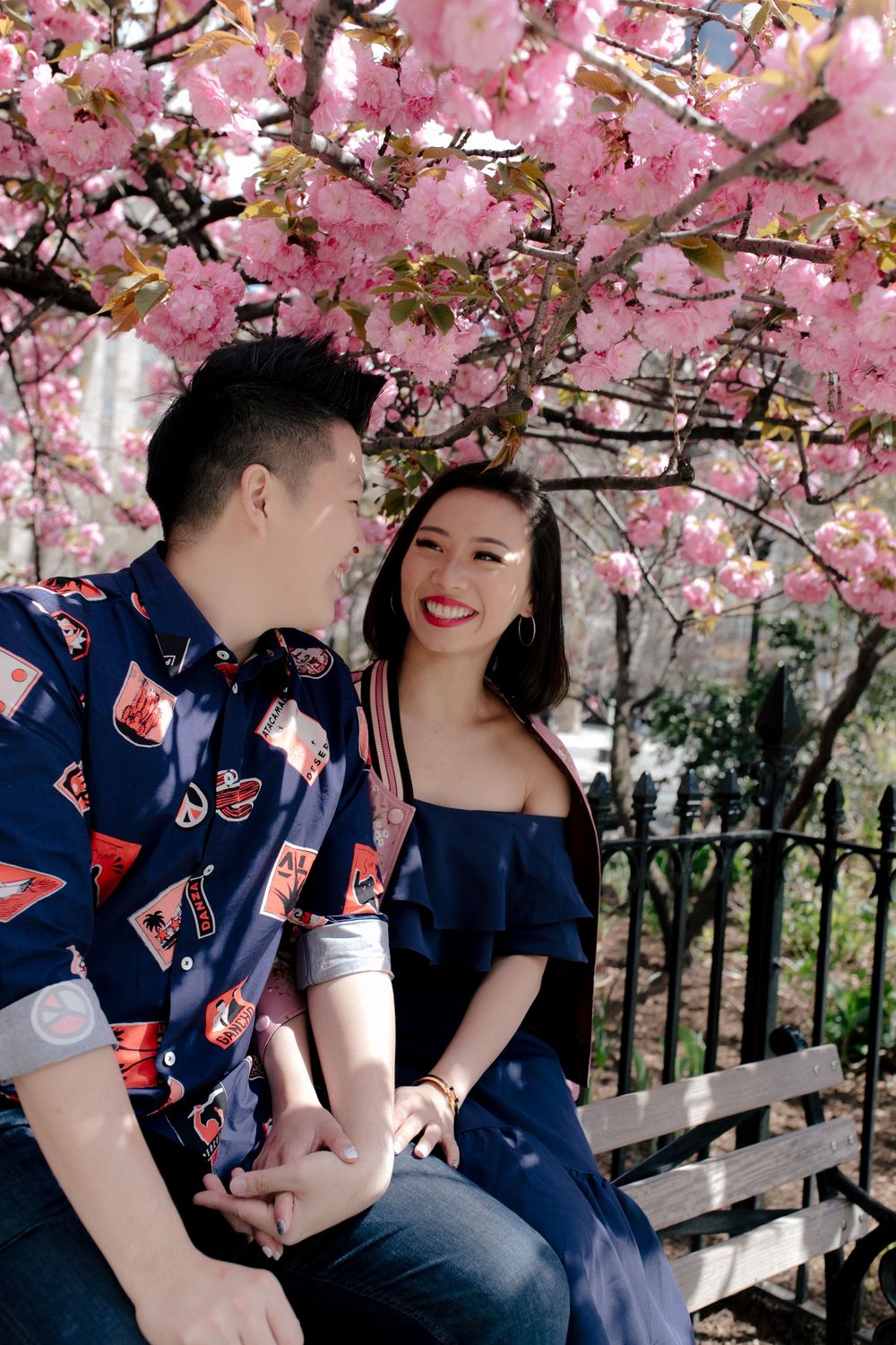 Couple sitting on bench under pink cherry blossom tree, holding hands and smiling at each other in spring.
