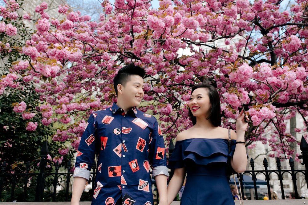 Asian couple holding hands under blooming pink cherry blossom trees, dressed in coordinating navy outfits.