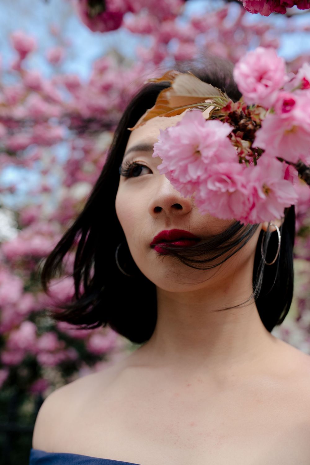 Asian woman with red lips posing among pink cherry blossom trees, flowers covering one eye.