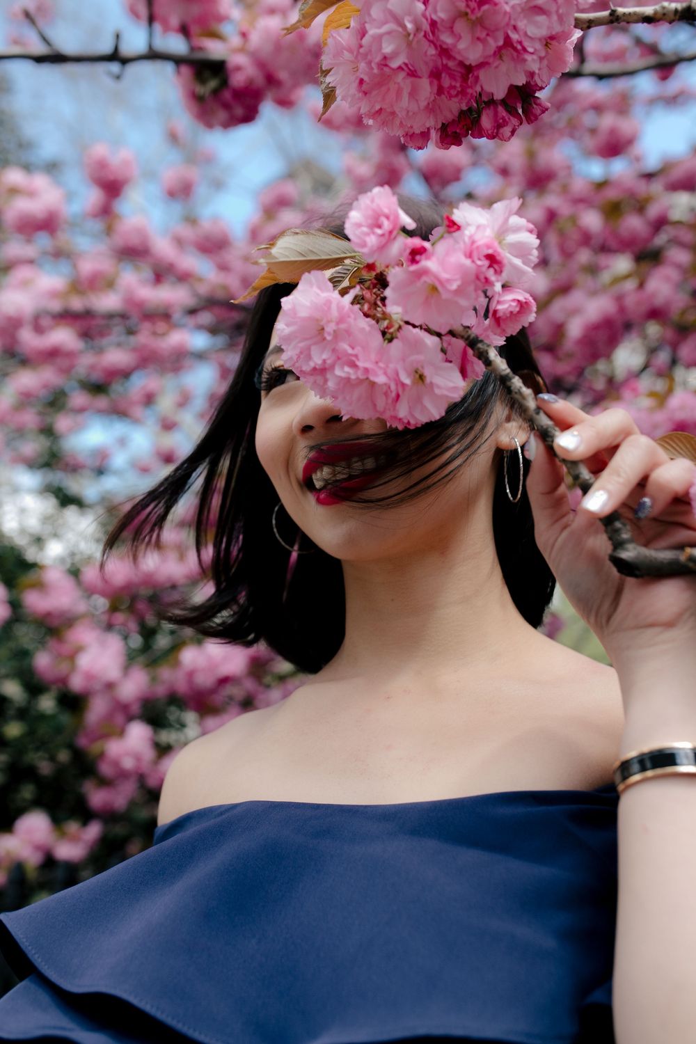 Woman in blue off-shoulder dress holding pink cherry blossom branch near her face, smiling outdoors.