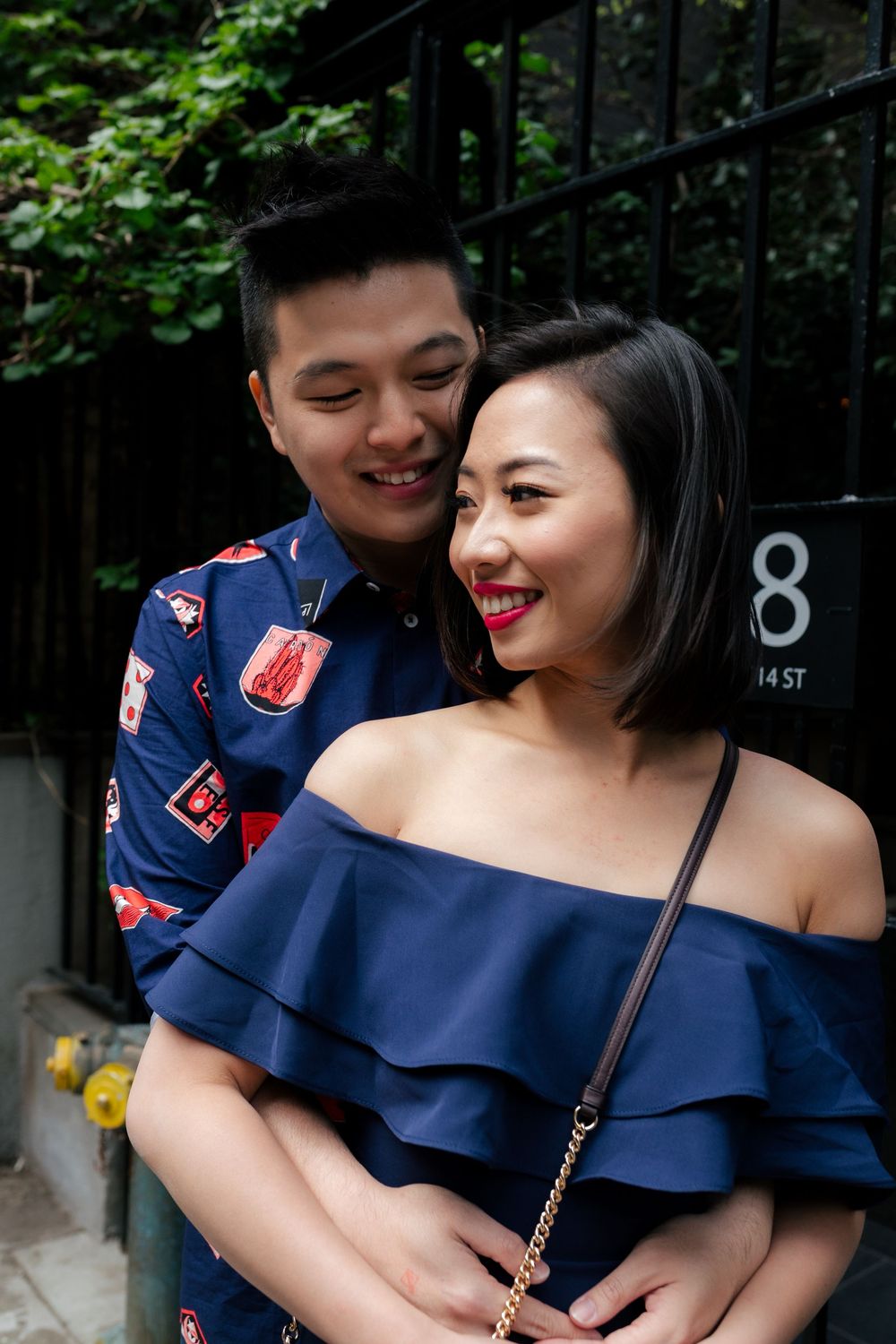 A smiling Asian couple embracing near a street sign, woman in navy off-shoulder dress, man in patterned shirt.