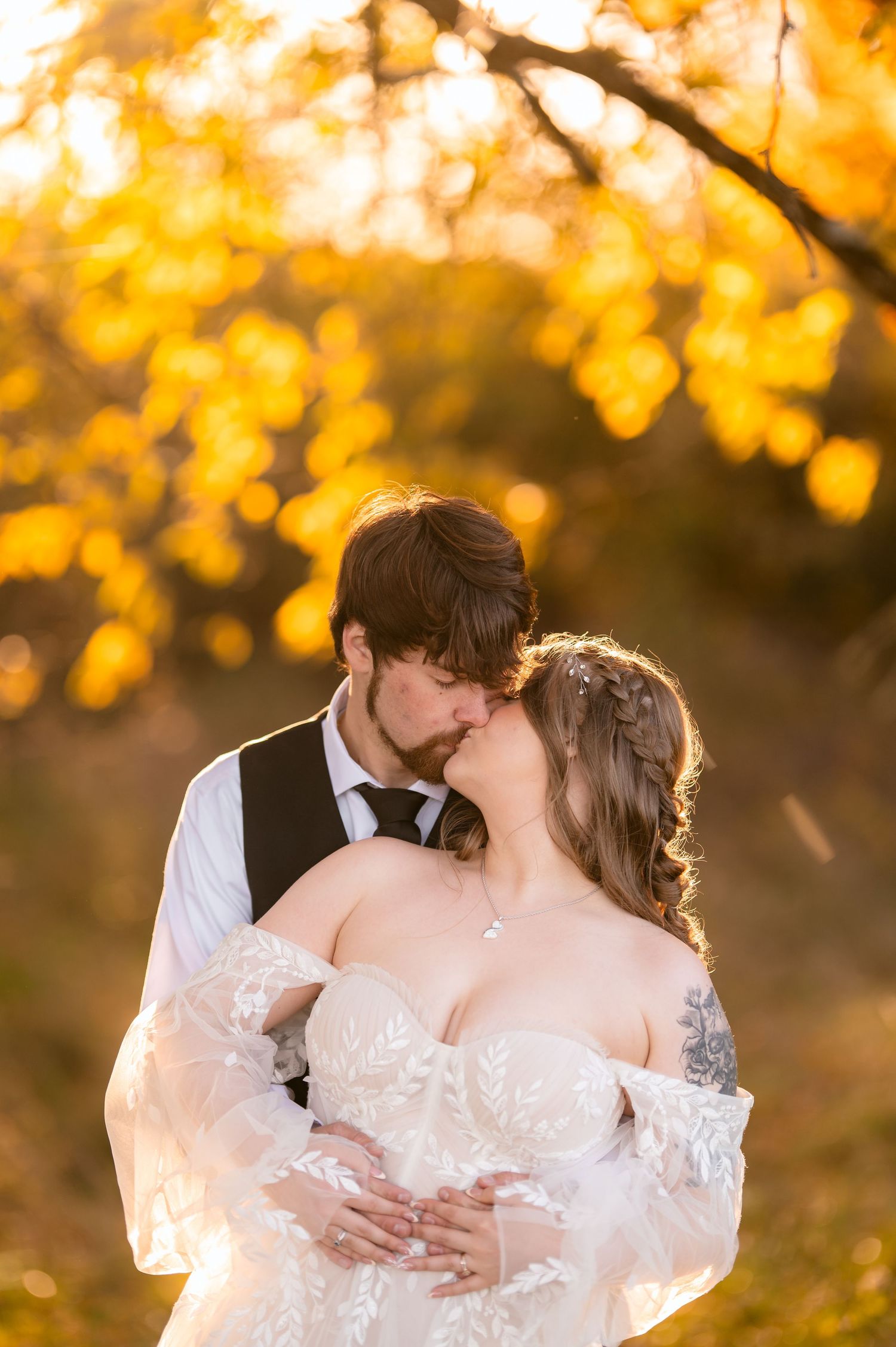 A bride and groom share a romantic kiss outdoors surrounded by golden autumn foliage during their wedding day.