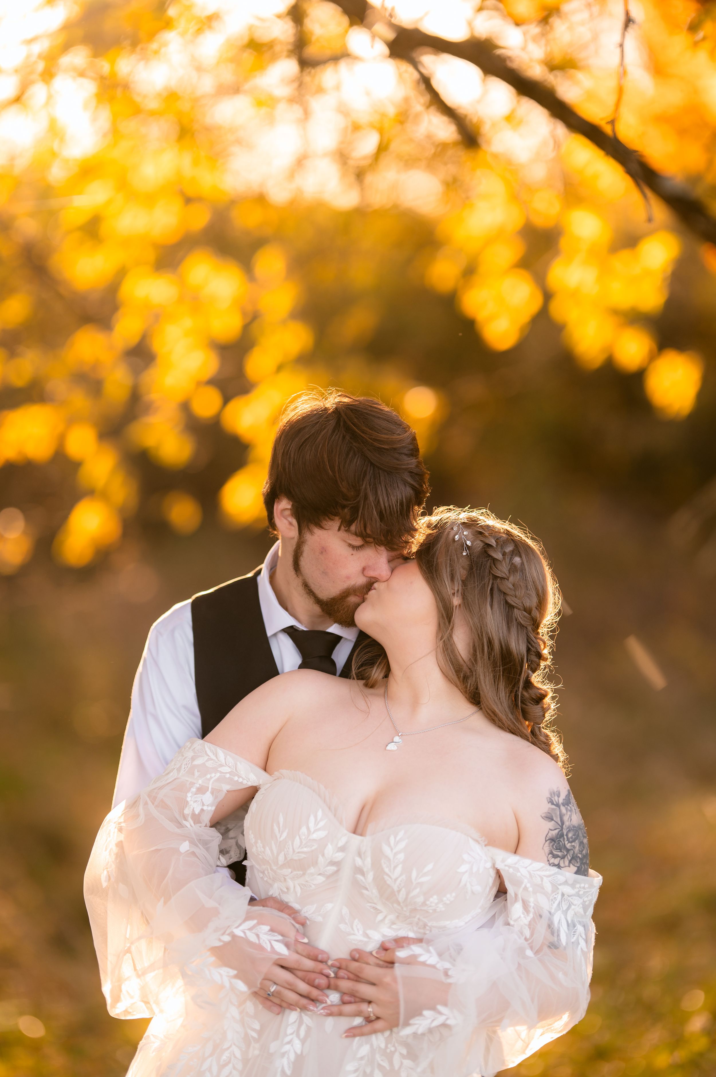 A bride and groom share a romantic kiss outdoors surrounded by golden autumn foliage during their wedding day.
