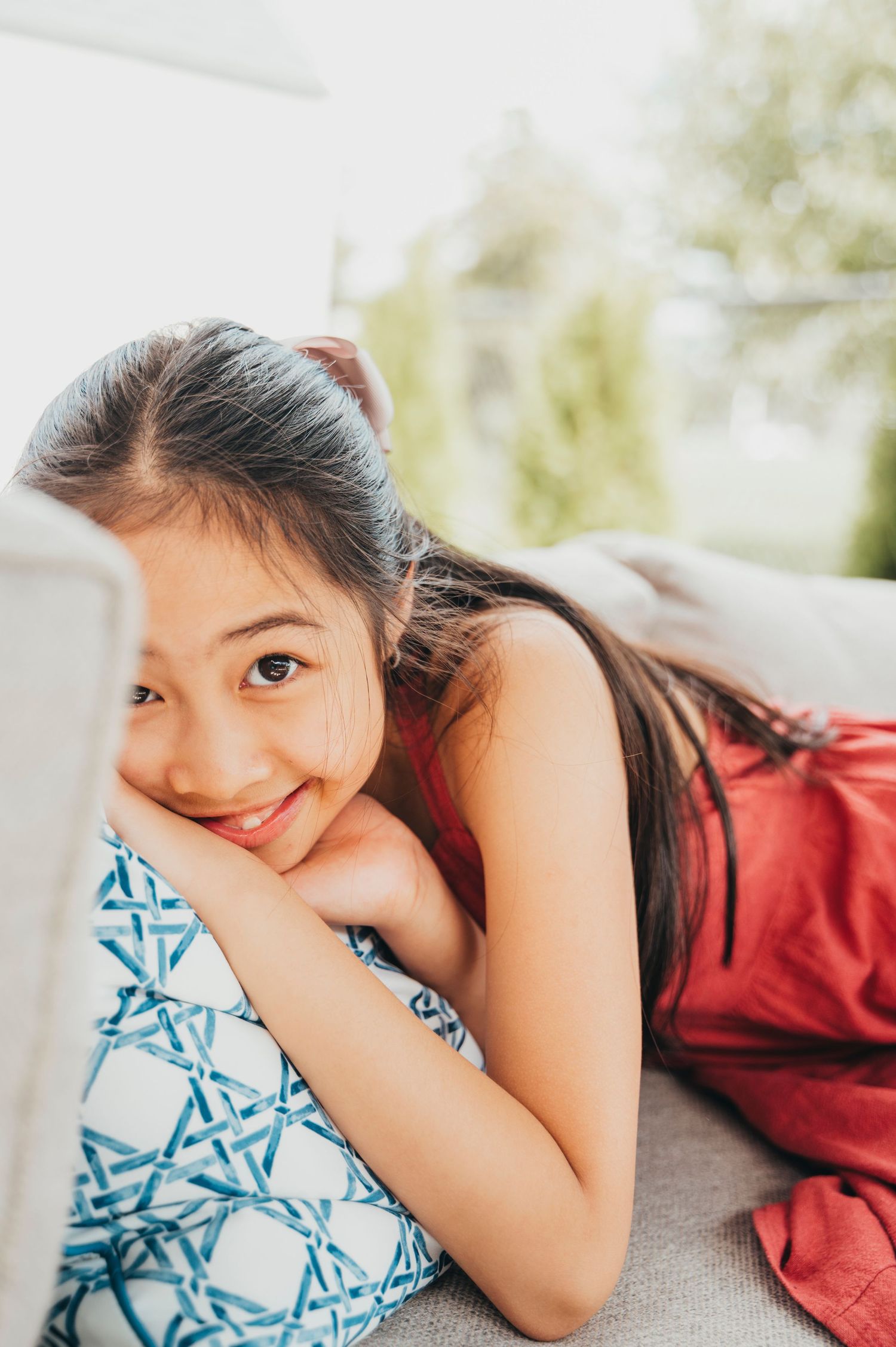 Little girl pops her face from behind arm of outdoor couch smiling.