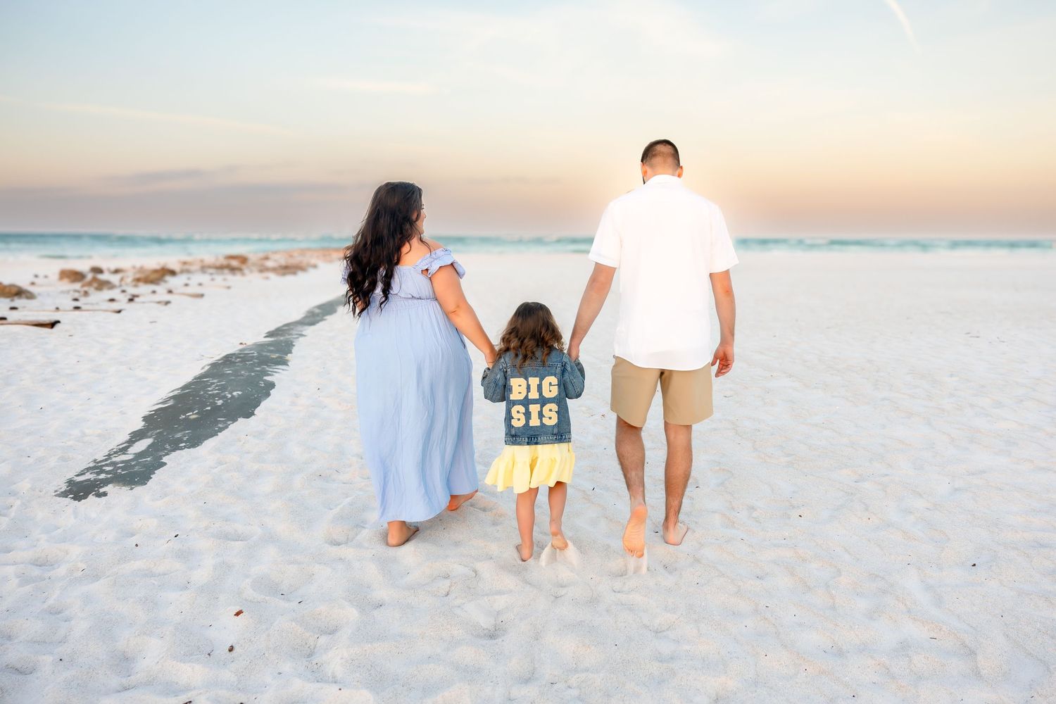 Family of three walking hand-in-hand on white sand beach at sunset, child wearing a Big Sis shirt.
