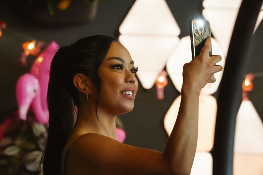 A young woman with a ponytail takes a selfie with her smartphone in a warmly lit, decorated venue.