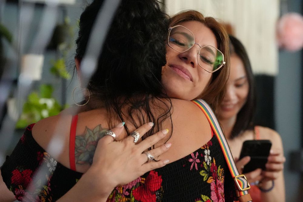 Two women sharing a warm hug at an outdoor social gathering with friends nearby.