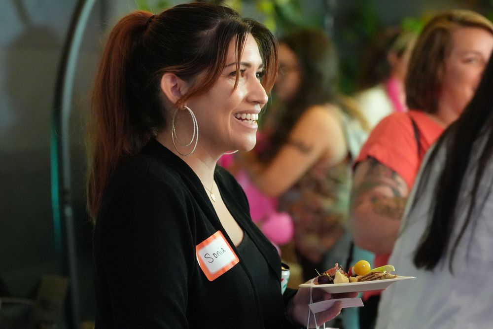 Smiling woman with hoop earrings holding a plate of food at a casual networking event.