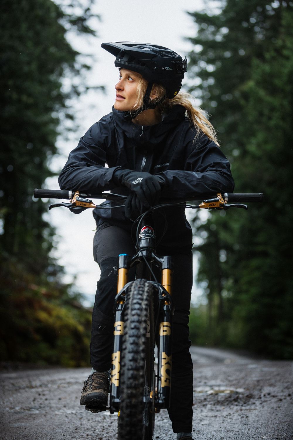 Woman mountain biker in black gear and helmet resting on handlebars on a forest trail.