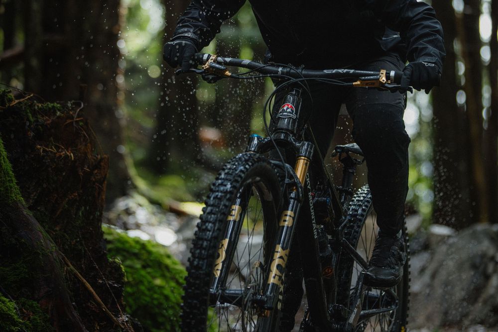 Mountain biker riding a muddy full-suspension bike through a lush green forest trail, wearing black gear.