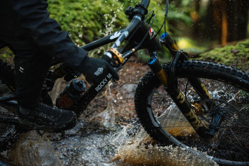 Mountain biker splashing through a muddy stream on a black full-suspension bike in a forest.